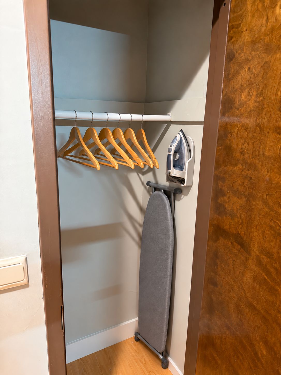 Un armario con perchas de madera, una tabla de planchar gris y un planchador fijado a la pared.