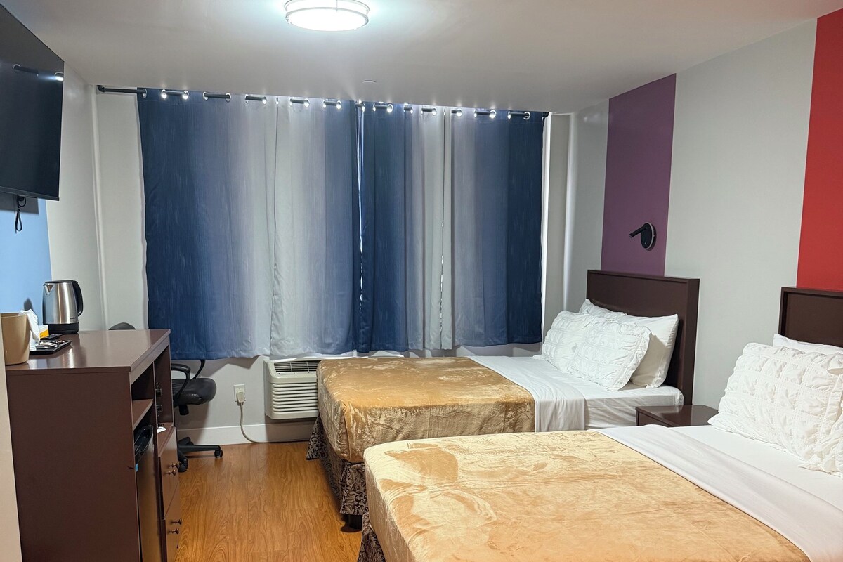 A hotel room with two beds, a desk with a kettle, a television, and a window with curtains.
