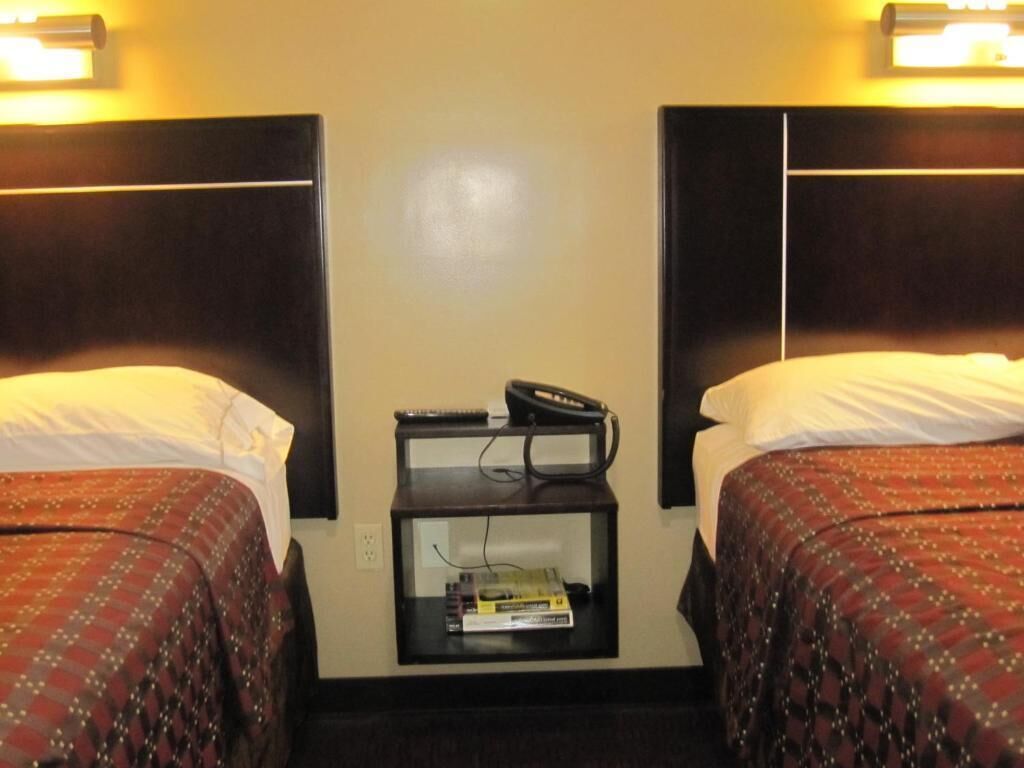 A hotel room with two beds, each with a headboard, a bedside table with a phone and books, and a wall-mounted light fixture.