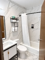 A bathroom with a toilet, sink, and bathtub. There is a shower curtain, a towel rack, and a small shelf with toiletries.