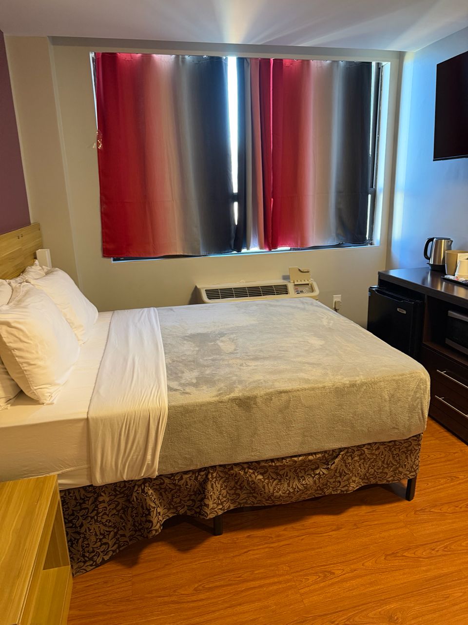 A hotel room with a bed, a bedside table, a heater, a window with curtains, and a small kitchenette area.