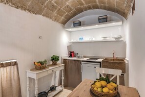 A compact kitchen with a brick archway, a white countertop, a small refrigerator, and a wooden dining table with a basket of lemons.