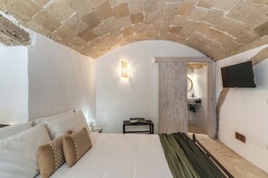 Ein Schlafzimmer mit Steindecke, einem Bett mit weißer Bettwäsche, einem Nachttisch, einem Flachbildfernseher und einem durch einen Bogen sichtbaren Badezimmer.