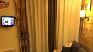 Un salon chaleureux avec un canapé en bois, une petite table sur laquelle se trouvent une bouteille et des verres, un téléviseur accroché au mur et une fenêtre avec des rideaux.