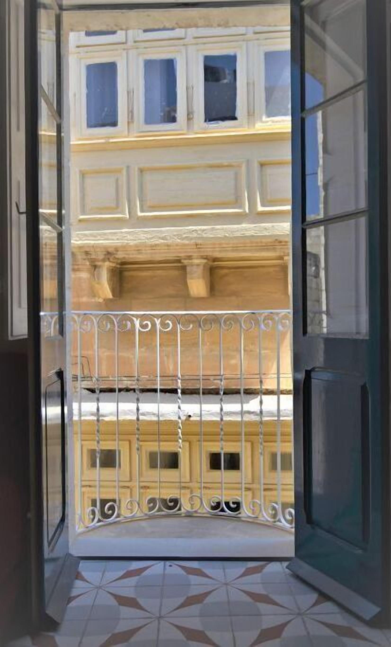 Vista a través d'una porta de vidre a un balcó amb barana metàl·lica, un sòl de rajoles i una façana d'edifici amb finestres i motllures decoratives.