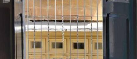 Vista a través d'una porta de vidre a un balcó amb barana metàl·lica, un sòl de rajoles i una façana d'edifici amb finestres i motllures decoratives.
