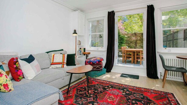 A living room with a sofa, a small table, a patterned rug, and a view of the outside. - Stunning 1BDR Basement Flat with Garden in Camden (London)