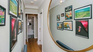 A hallway with framed artwork on the walls, a round mirror, and a bench. - Stunning 1BDR Basement Flat with Garden in Camden (London)