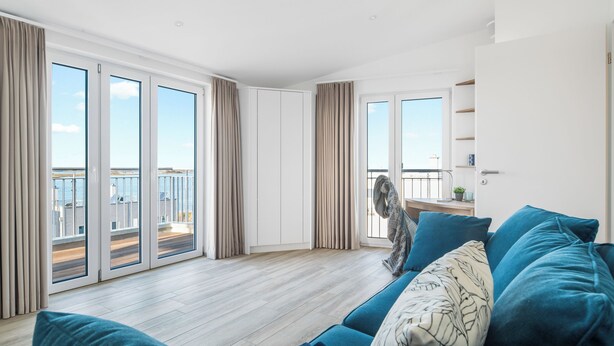 A modern living room with a blue sofa, a fan, and a view of a balcony with a railing. - Admiralsdeck Panoramablick, Finnische Sauna, 3 Baeder, Fahrstuhl, 2 Terrassen (Olpenitz)
