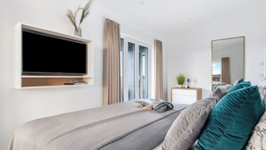 A modern bedroom with a large bed, a TV mounted on the wall, a bedside table with a lamp, and a window with curtains. - Admiralsdeck Panoramablick, Finnische Sauna, 3 Baeder, Fahrstuhl, 2 Terrassen (Olpenitz)