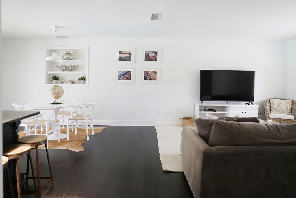 A modern living room with a white sofa, a dining area with a white table and chairs, a flat-screen TV on a white stand, and a small shelf with decorative items.