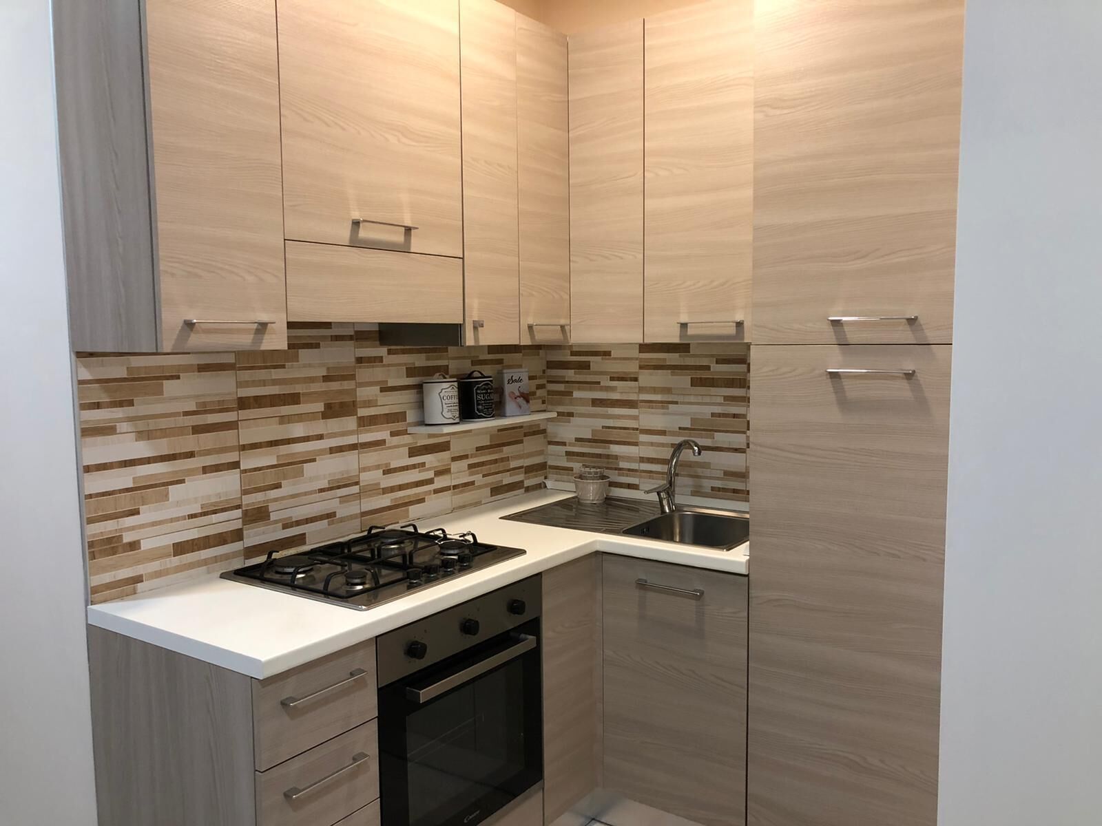 Een kleine keuken met een gasfornuis, oven en spoelbak. Er zijn houten keukenkastjes en een betegelde achterwand.