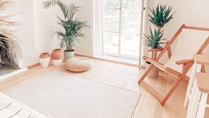 Quarto amplo e iluminado, com piso de madeira, uma grande janela com cortinas, luminária pendente e vários vasos com plantas.