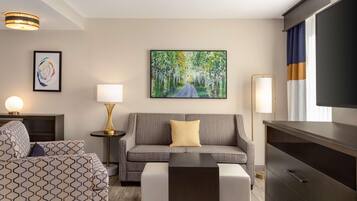 A modern hotel room with a grey sofa, a patterned bed, a flat-screen TV, and a painting of a forest path.