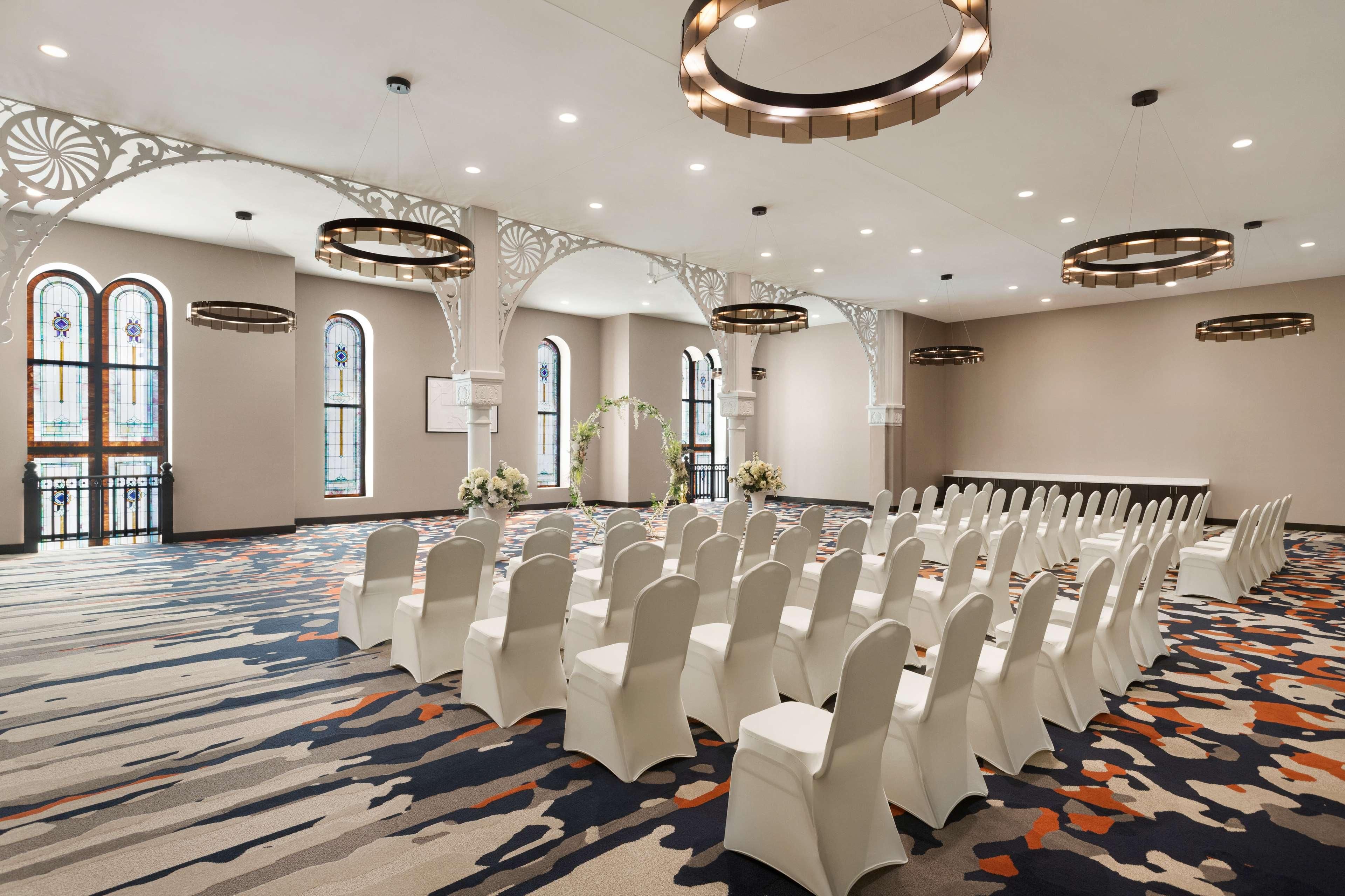 A banquet hall with rows of white chairs, large windows with stained glass, and decorative ceiling lights.