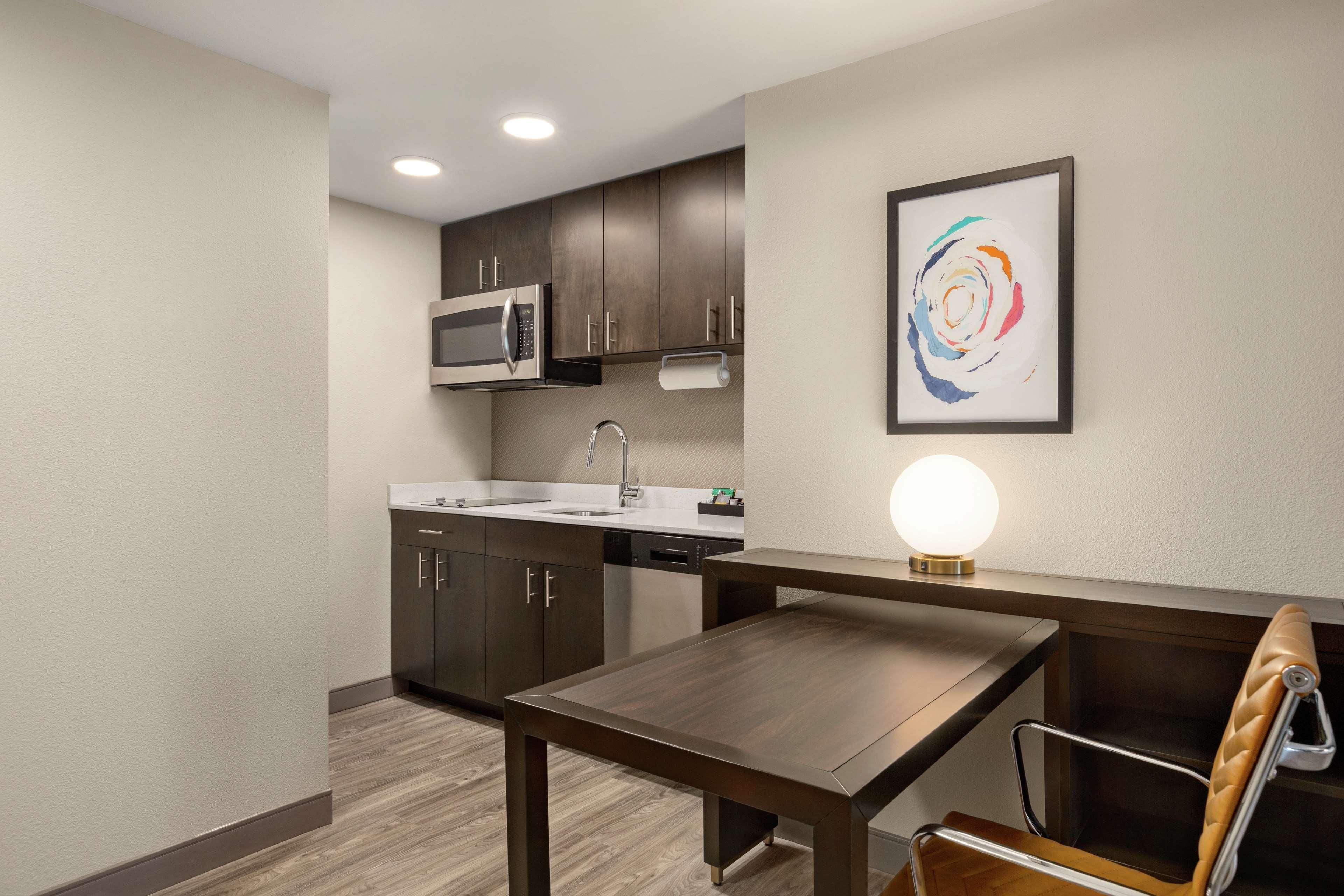 A modern kitchen with dark cabinetry, a stainless steel microwave, and a sink. There's a dining table with a chair and a wall-mounted light fixture.