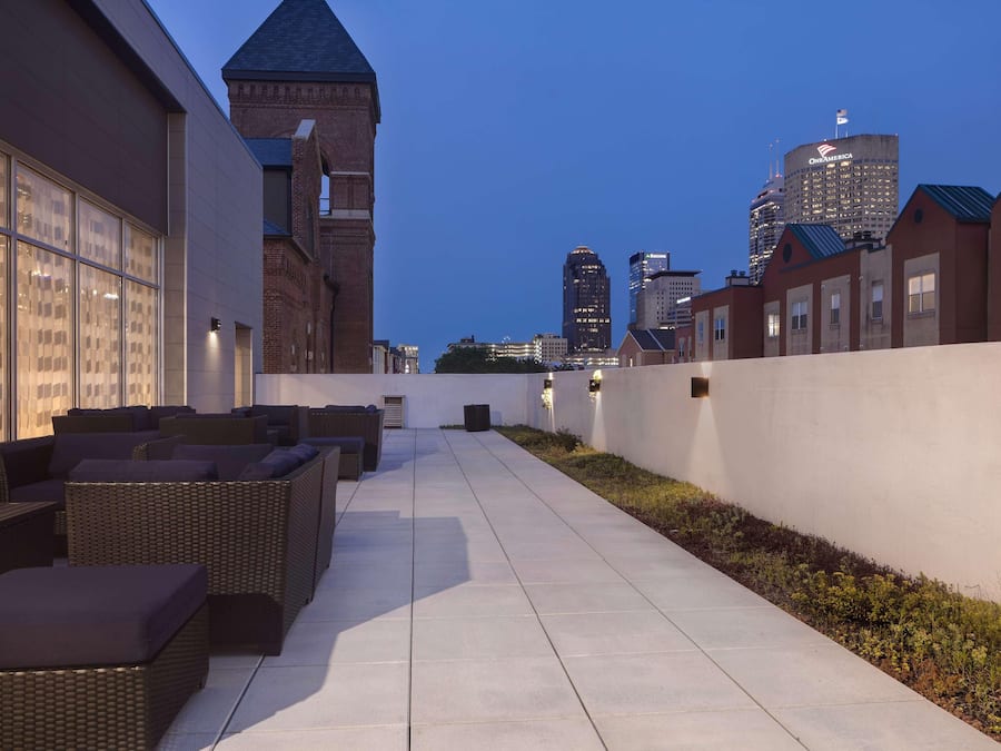 Una terraza en la azotea con mobiliario exterior, el horizonte de una ciudad al fondo y un edificio de ladrillo con torre.