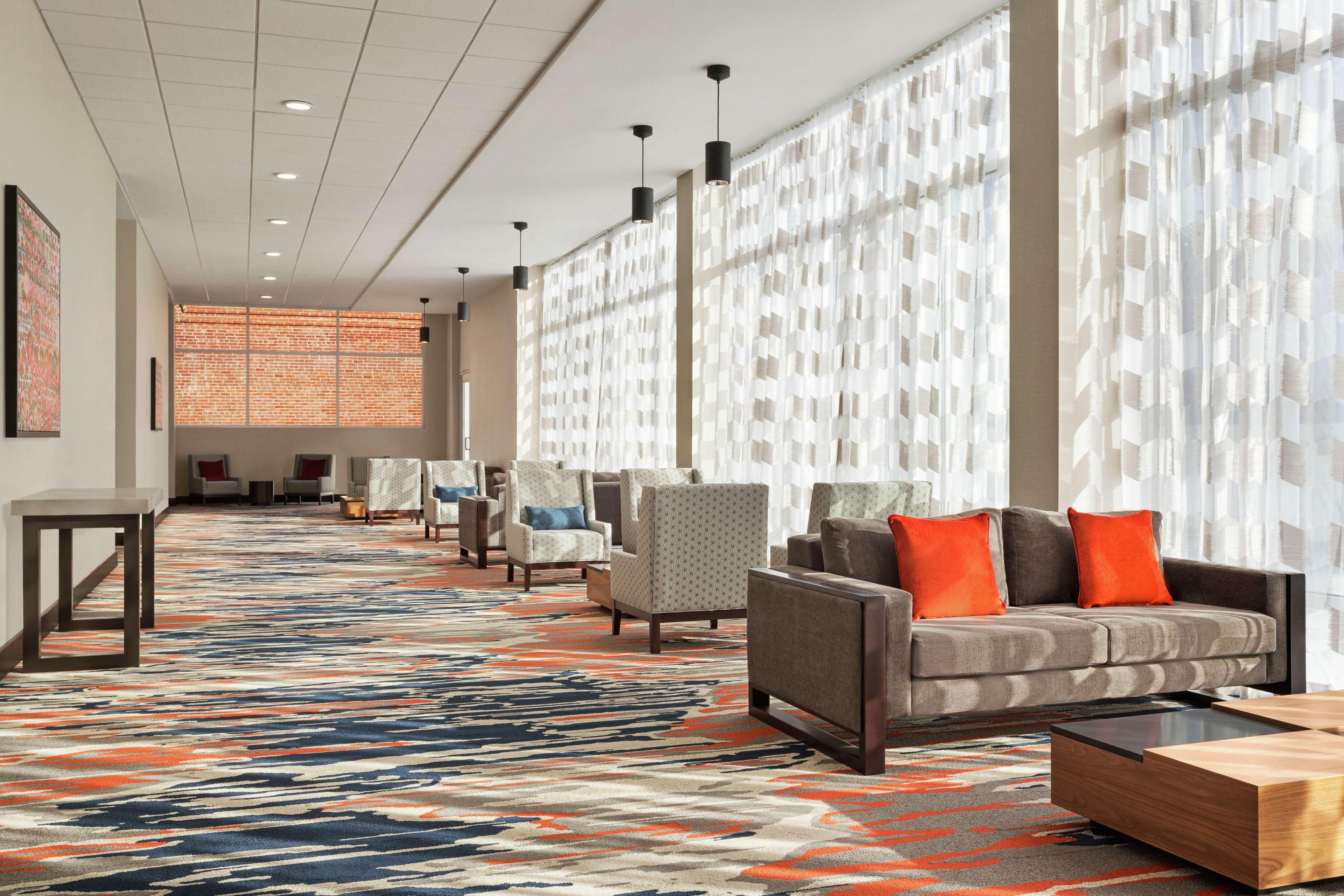 A hotel lobby with a patterned carpet, seating area with sofas and chairs, and large windows with sheer curtains.