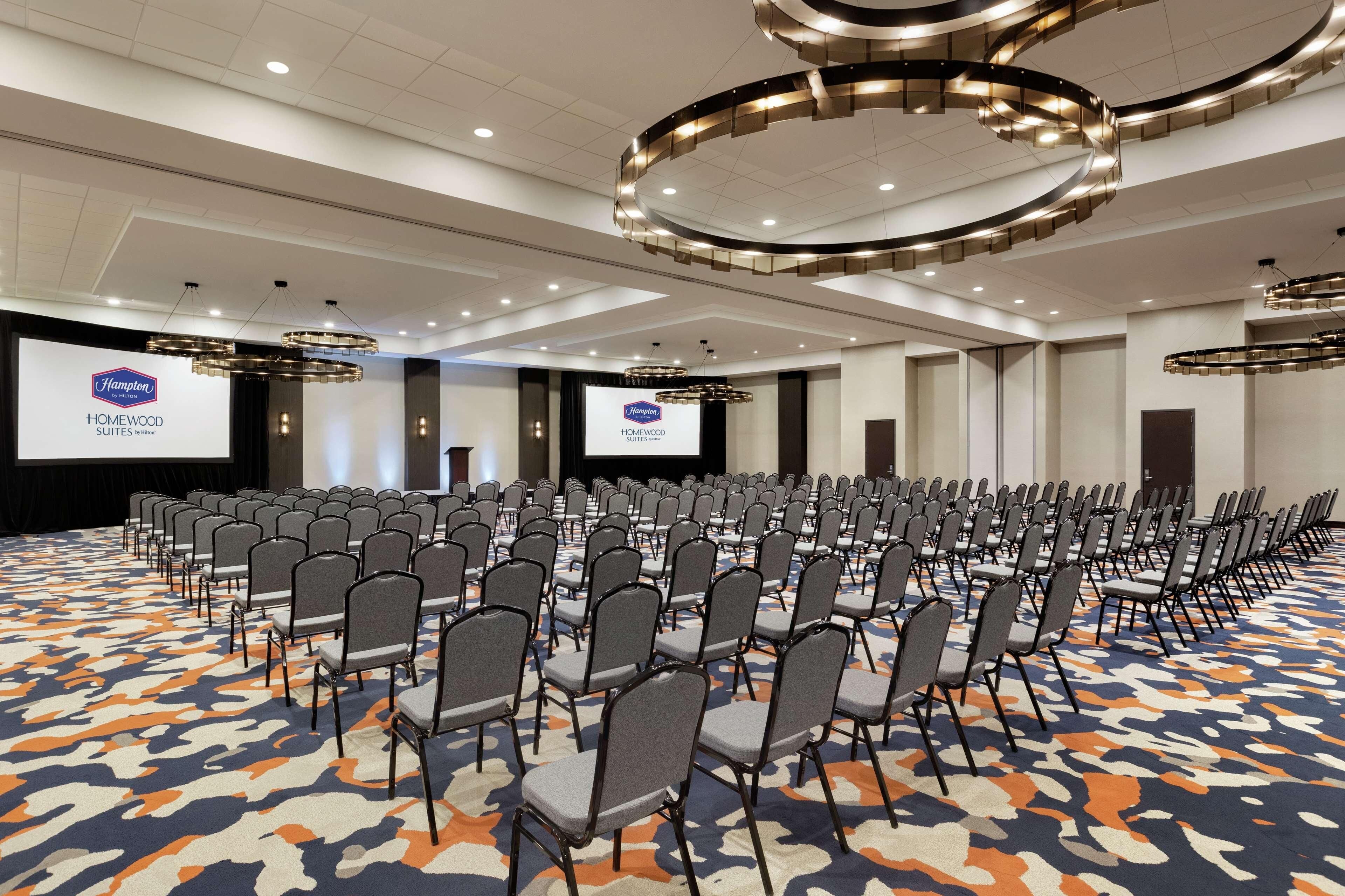 A conference room with rows of chairs, a large screen, and a logo for Hampton Homewood Suites.