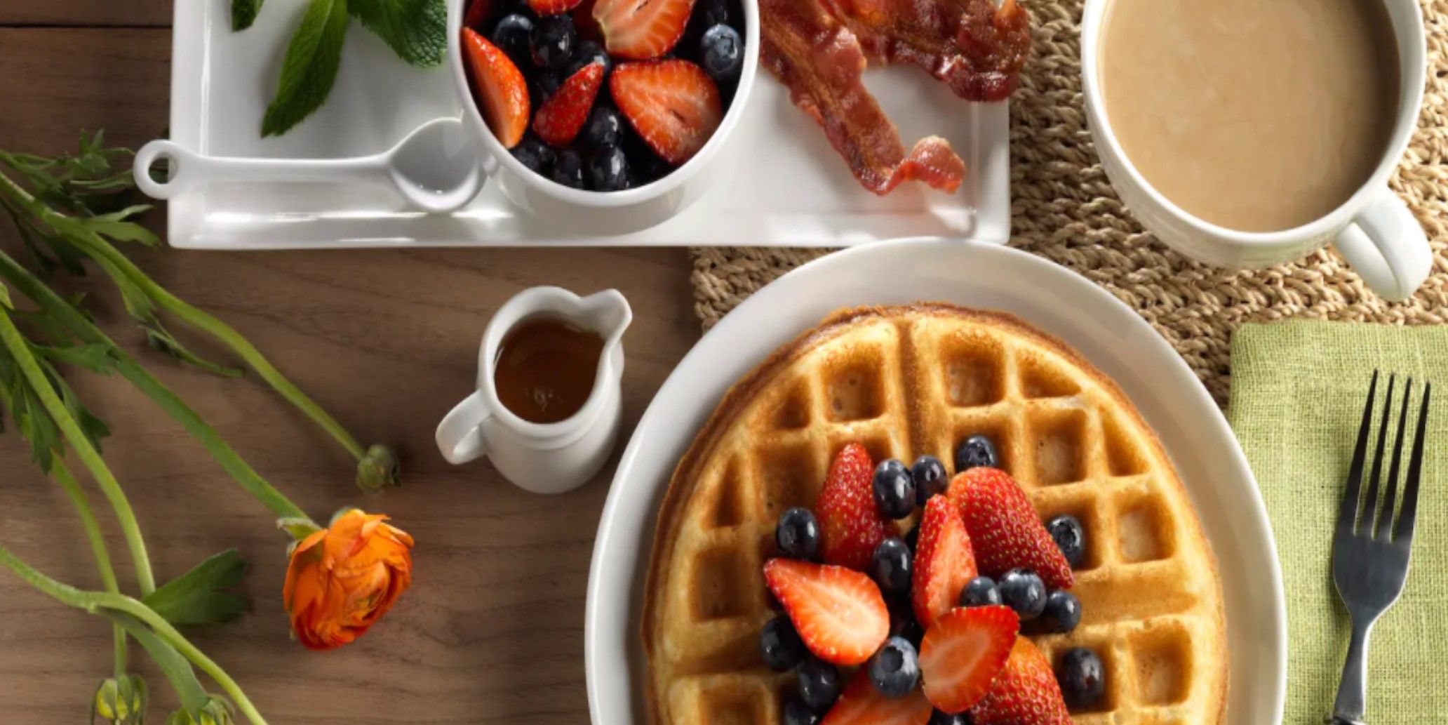 Un desayuno con waffles cubiertos de fresas y arándanos, un tazón pequeño de frutos rojos, un trozo de tocino y una taza de café.