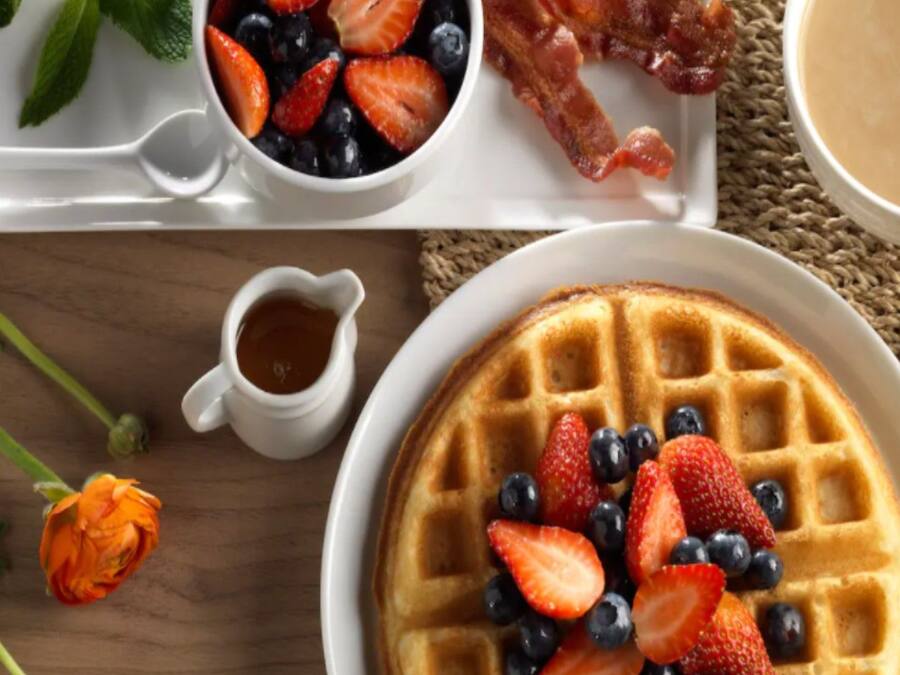 A breakfast setting with waffles topped with strawberries and blueberries, a small bowl of mixed berries, a piece of bacon, and a cup of coffee.