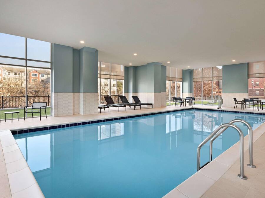 A swimming pool with lounge chairs and tables, large windows, and a view of buildings outside.