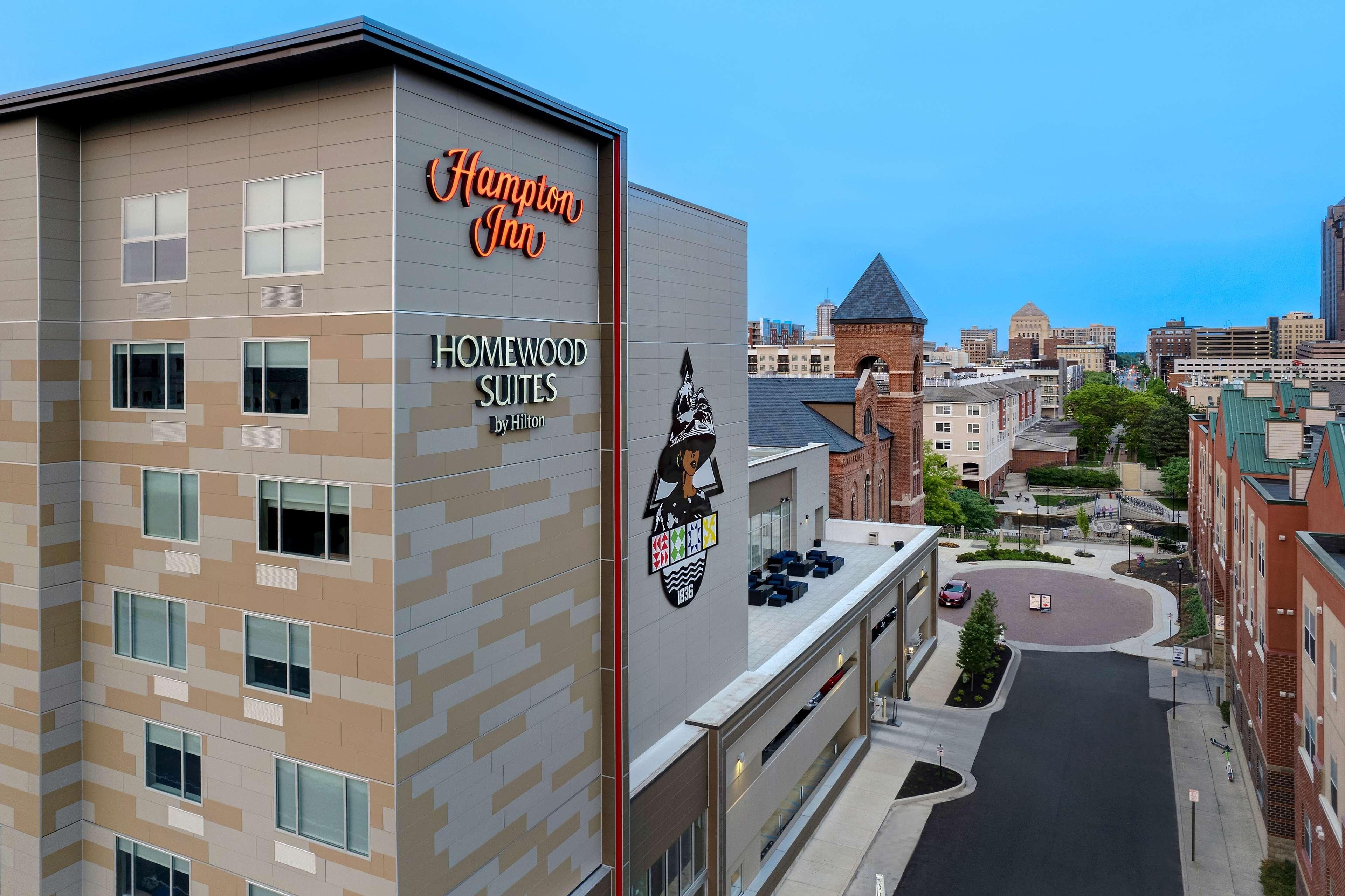 A Hampton Inn hotel with Homewood Suites by Hilton, featuring a modern design with a mix of beige and gray tones, multiple windows, and a prominent logo.