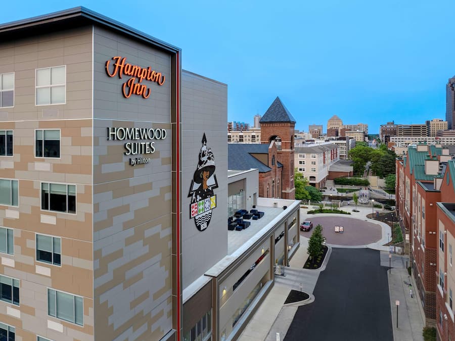 Um hotel Hampton Inn ao lado de um Homewood Suites by Hilton, com design moderno, misturando tons de bege e cinza, várias janelas e um logotipo bem visível.