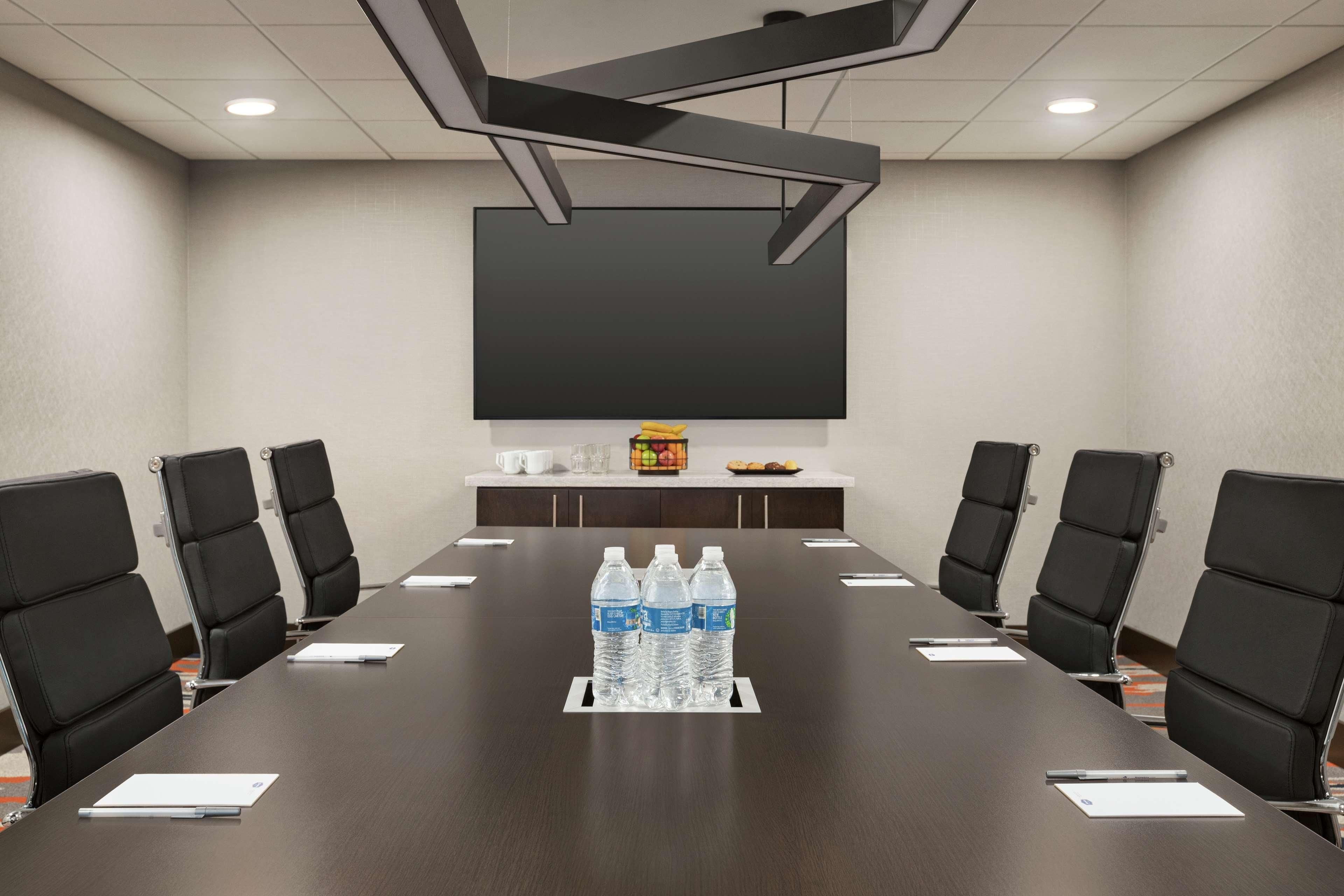 Un salón de conferencias con una larga mesa, sillas negras, botellas de agua y una pantalla de proyector.