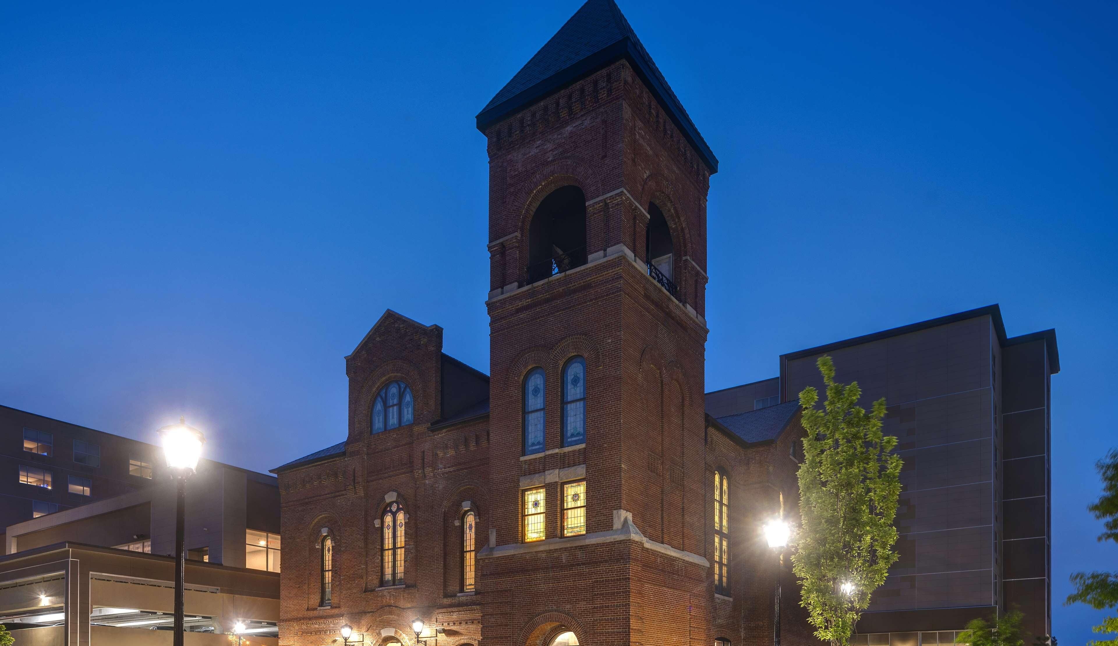 A historic brick building with a prominent tower and arched windows, surrounded by modern structures.