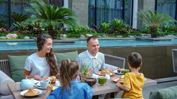 Une famille prend son repas à l’extérieur, près d’une piscine, avec une végétation luxuriante en arrière-plan.