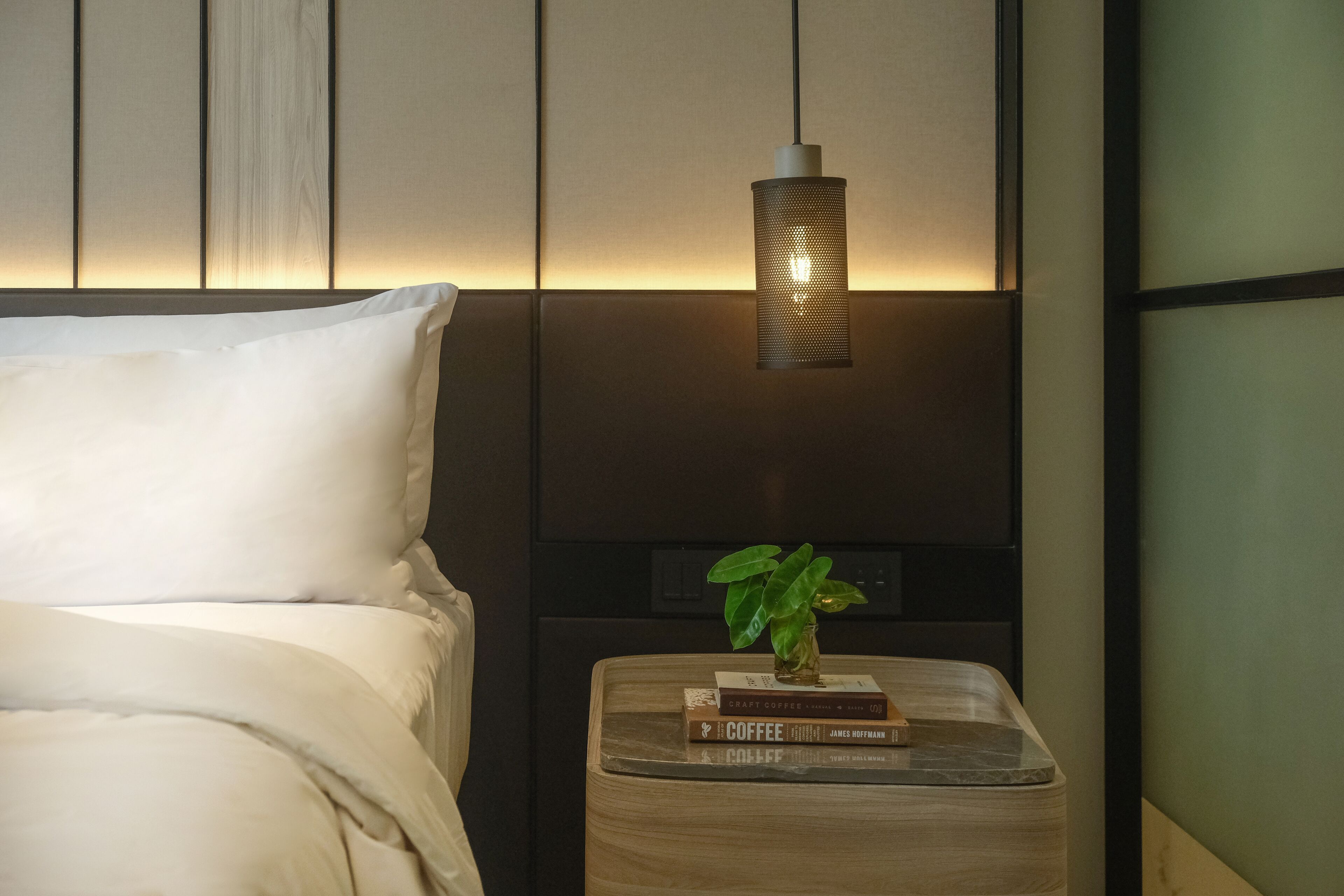 A neatly made bed with white linens, a bedside table with a plant and a stack of books, and a hanging light fixture.