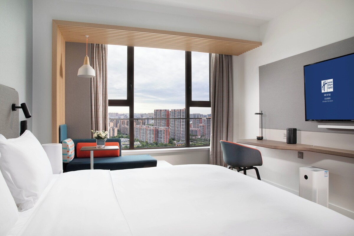 Ett hotellrum med en stor säng, ett skrivbord, en stol, en tv och utsikt över stadsbilden.
