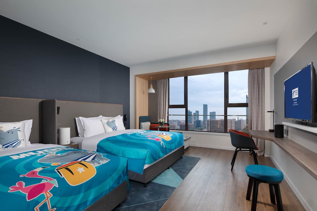 A modern hotel room with two beds, a desk, and a large window offering a city view.
