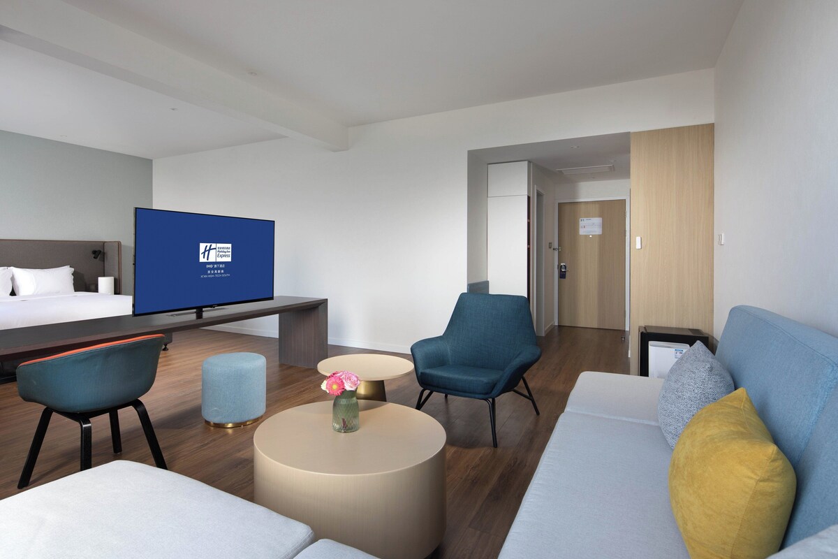 Ett hotellrum med en säng, en tv, ett skrivbord, en soffa, en stol, ett litet bord med en vas och en dörr som leder till ett annat rum.