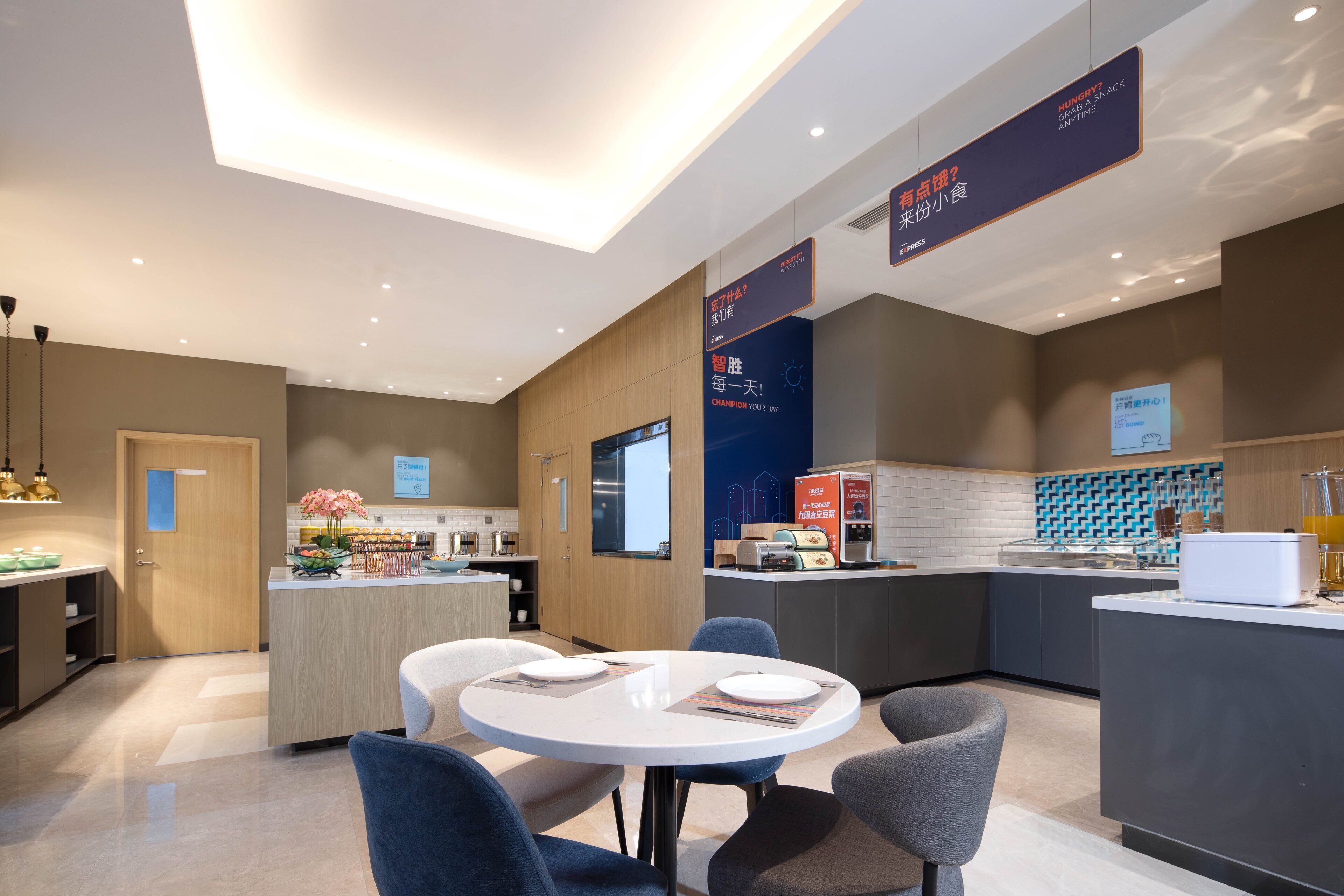 A modern hotel lobby with a round dining table, comfortable seating, a reception desk, and a kitchen area in the background.