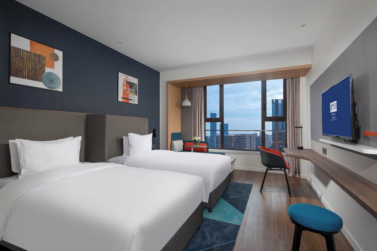 Ett hotellrum med två sängar, ett skrivbord, en stol, en TV och ett stort fönster med utsikt över staden.