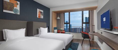 A hotel room with two beds, a desk, a chair, a TV, and a large window with a city view.