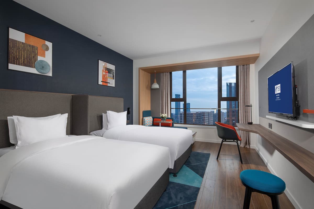 A hotel room with two beds, a desk, a chair, a TV, and a large window with a city view.
