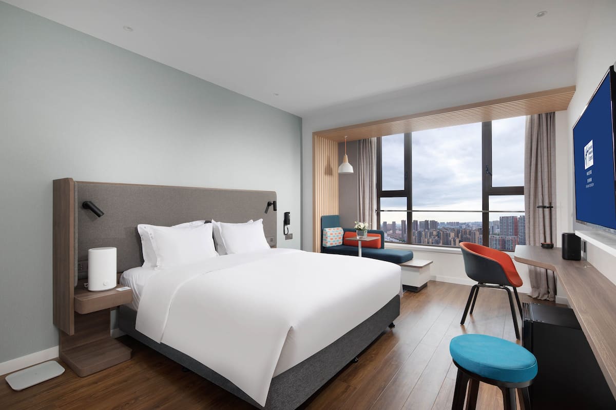 A modern hotel room with a large bed, a desk, a chair, and a view of the city.