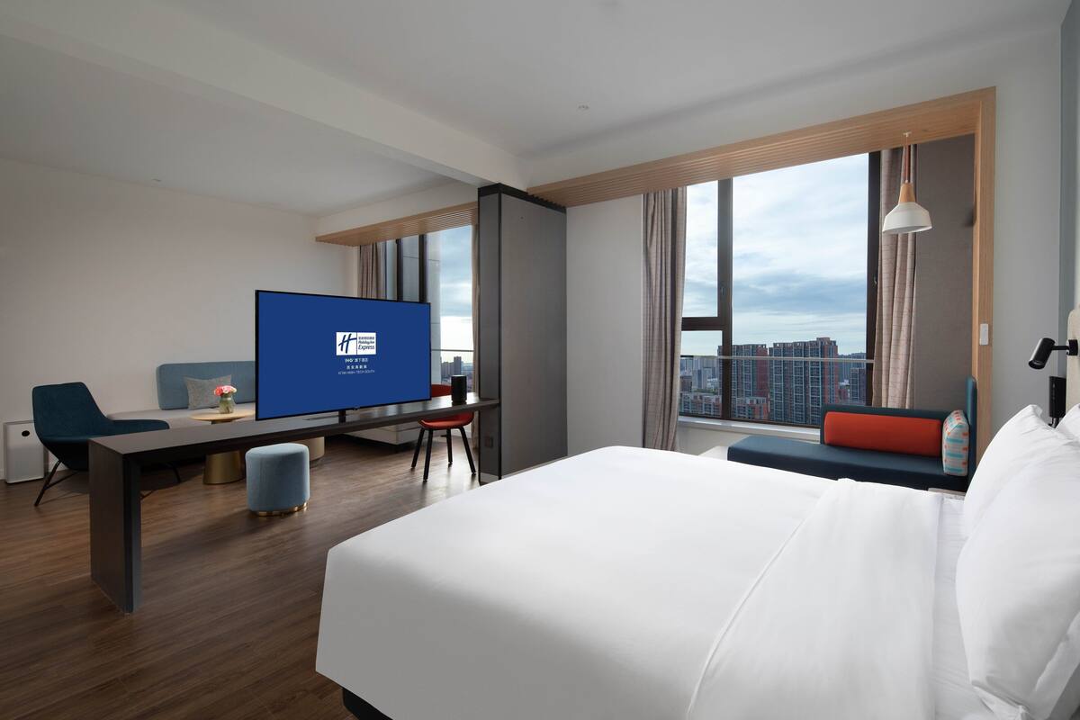 A modern hotel room with a large bed, a desk with a TV, a seating area with a view of the city, and large windows.