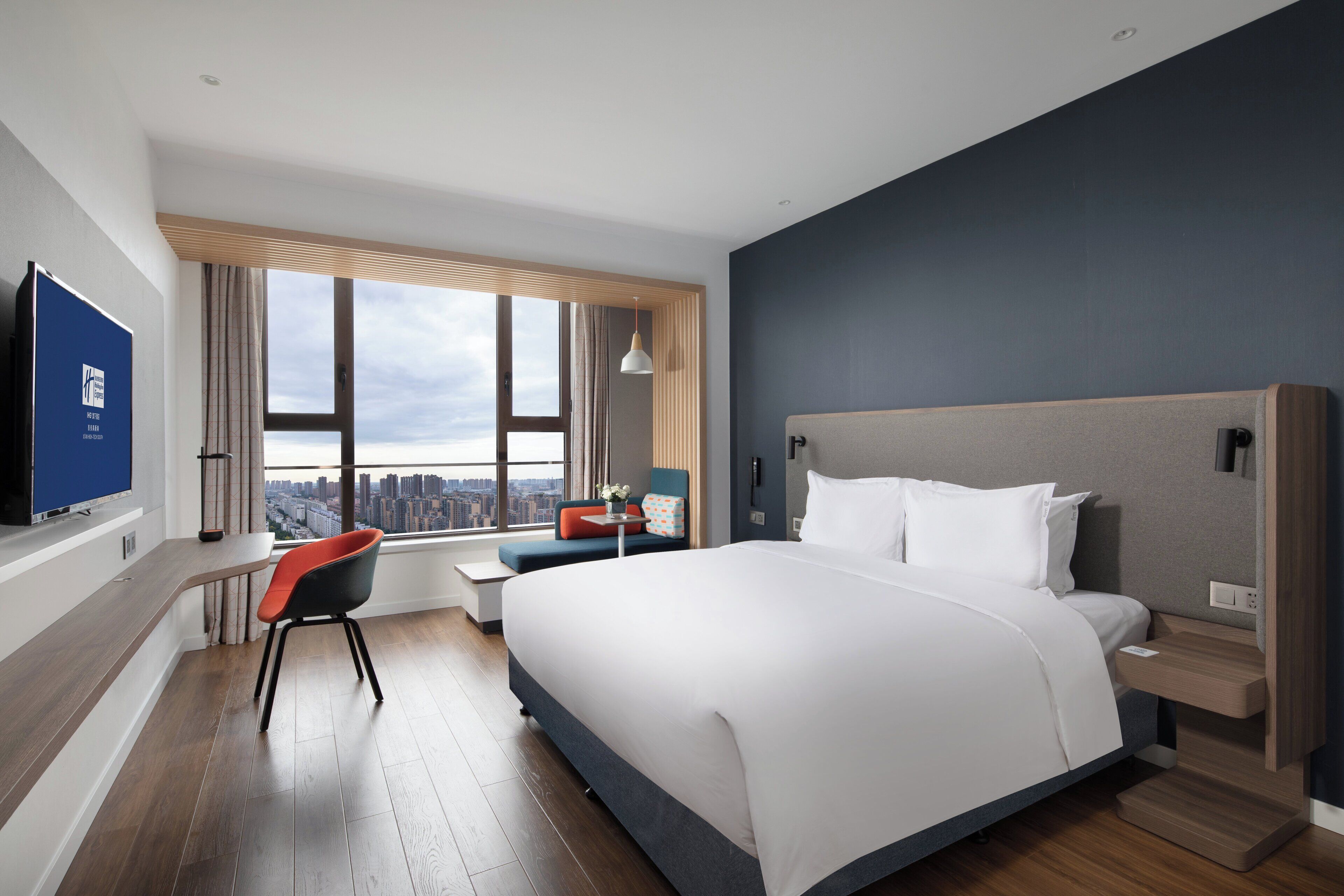 A modern hotel room with a large bed, a desk, and a chair. There is a view of the city through the window.