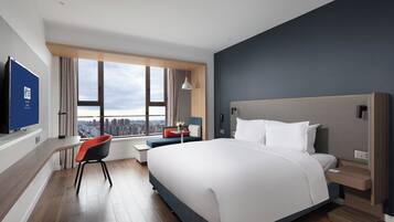 A modern hotel room with a large bed, a desk, and a chair. There is a view of the city through the window.