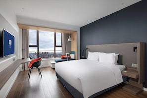 Une chambre d’hôtel moderne avec un grand lit, un bureau et une chaise. La fenêtre offre une vue sur la ville.