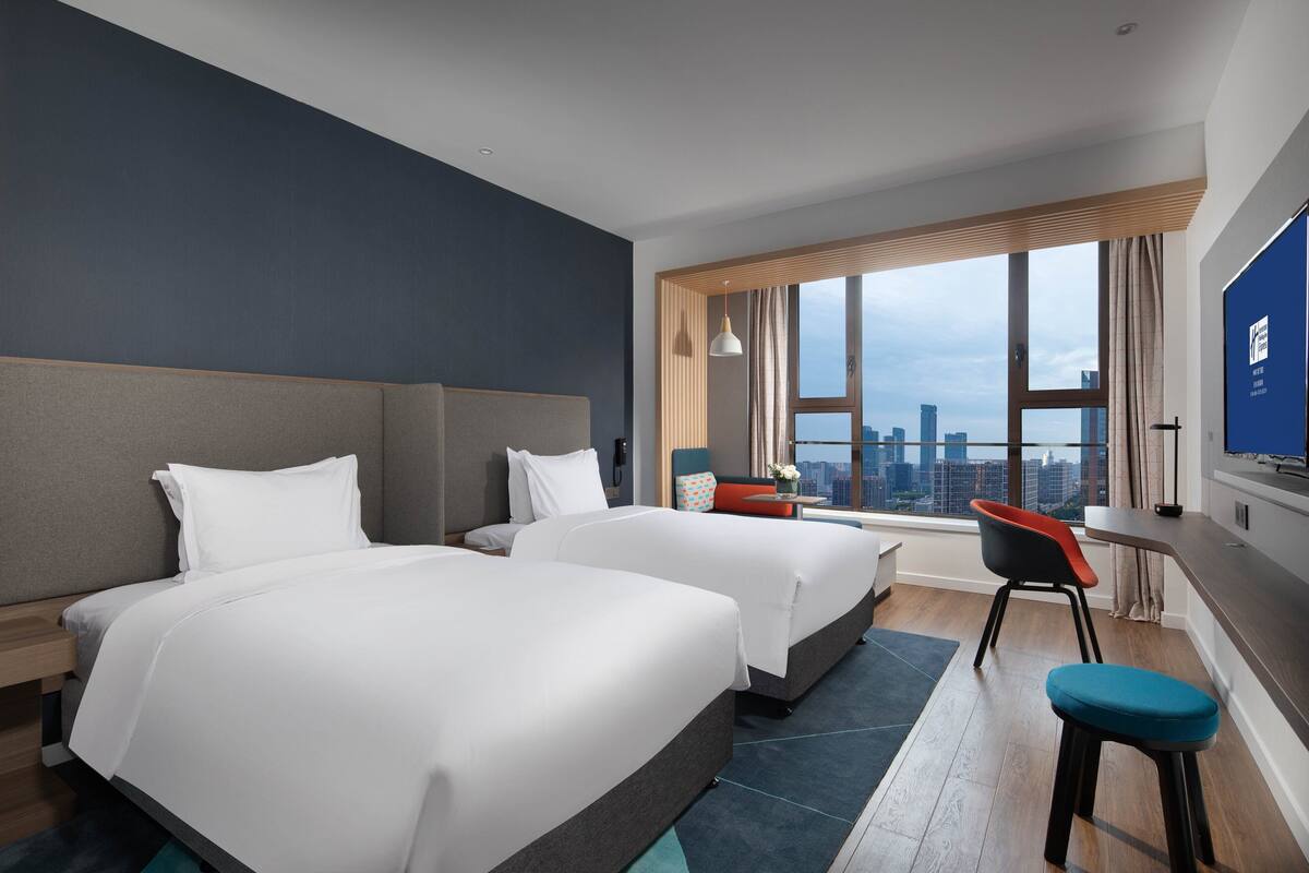 A hotel room with two beds, a desk, a chair, and a large window with a city view.