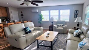 Ein Wohnzimmer mit einem beige gefärbten Sofa, einem Holztisch und einem Deckenventilator. Es gibt eine Küchenzeile mit Holzschränken und einer Arbeitsplatte.
