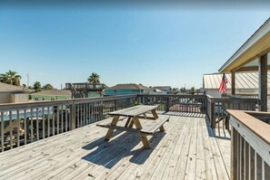 Une terrasse en bois avec une table de pique-nique, une rambarde et une vue sur la plage et les bâtiments en arrière-plan.