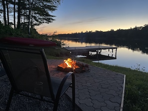 A lakeside patio with a fire pit, a dock, and a chair.