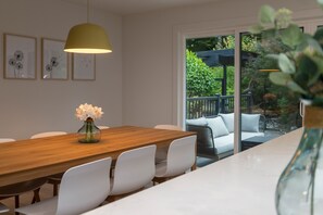 Uma sala de jantar moderna com uma mesa de madeira e cadeiras brancas, um vaso com flores e vista para um pátio externo com um sofá branco e uma pérgula.
