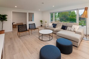 Uma sala de estar moderna com um sofá, dois pufes, uma mesa de centro e uma poltrona. Há uma janela grande com vista para o verde lá fora.